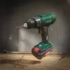 A cordless drill is shown in a dynamic pose, with drill bits and wood shavings scattered around it, against a textured concrete background. Produktbild-Vorschau