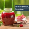 A jar of red jam is being held by a green jar lifting tool, with raspberries and mint leaves in the foreground. Produktbild-Vorschau