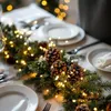 A festive table setting with a garland of pine branches and fairy lights, decorated with pinecones, set for a meal. Voir les images du produit