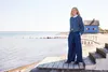 A woman stands on a wooden deck by the sea, with a blue house in the background. Produktbild-Vorschau
