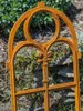 A close-up of a rusty metal garden trellis with a fleur-de-lis design, set against a backdrop of green plants and stones. Produktbild-Vorschau