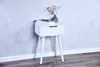 A white bedside table with a drawer and three legs, placed against a textured gray wall. A book and a vase with flowers are on top of the table. Produktbild-Vorschau