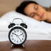 A retro alarm clock sits on a wooden surface in front of a person sleeping in bed. Voir les images du produit