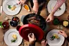 A group of people are gathered around a table, serving a roasted chicken from a large pot. Produktbild-Vorschau