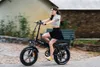 A woman is riding a black electric bicycle on a paved path, with a building and trees in the background. Anteprima dell’immagine prodotto