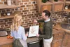 A couple is in a kitchen, with the man holding a cutting board and the woman cooking. Produktbild-Vorschau