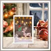 A Christmas-themed scene with a framed picture of a reindeer, snowman, and Christmas decorations, set against a backdrop of a Christmas tree and a snowy window. Anteprima dell’immagine prodotto