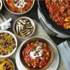A spread of food dishes, including a slow cooker filled with stew, couscous, grilled eggplant, and a bowl of stew with yogurt and pomegranate seeds. Produktbild-Vorschau