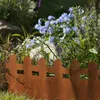 A rusty metal garden border with a picket fence design, separating a grassy area from a flower bed with blue flowers and green foliage. Produktbild-Vorschau