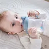 A baby lying on a white blanket, drinking from a sippy cup. Produktbild-Vorschau