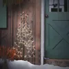 A lighted Christmas tree decoration is placed outside a wooden wall next to a green door, with snow on the ground. Podgląd zdjęcia produktu