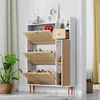 A white shoe cabinet with rattan-fronted flip-down doors and a small drawer, placed in a room with a plant and curtains. Produktbild-Vorschau