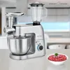 A silver multi-functional kitchen appliance with a mixer bowl, blender attachment, and meat grinder attachment, placed on a countertop in a kitchen setting. A bowl of ground meat is in the foreground. Produktbild-Vorschau