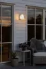 An outdoor wall light is illuminated on the side of a white house with large windows. A chair with a furry throw is visible to the right. Produktbild-Vorschau