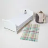 A white baby bed with a white mattress protector and a small stuffed animal on it, next to a gray pouf with blankets and a plaid rug on a white background. Produktbild-Vorschau