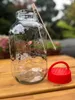 A glass jar with a red lid and a wooden stick on a wooden table, with a blurred garden background. Produktbild-Vorschau