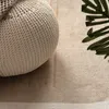 A close-up view of a textured beige pouf resting on a beige rug with a hint of a green leaf in the corner. Anteprima dell’immagine prodotto