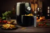 An air fryer filled with french fries sits on a wooden table, surrounded by fresh ingredients and cooked food. Podgląd zdjęcia produktu