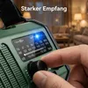A hand is shown tuning a green emergency radio in a living room setting. Produktbild-Vorschau
