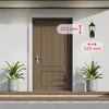 A classic outdoor wall lamp is shown mounted on a white wall next to a brown wooden door. Two potted plants flank the door on a wooden step. Produktbild-Vorschau