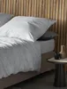 A close-up view of a bed with grey bedding and a wooden headboard. Produktbild-Vorschau