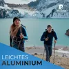 Two hikers with backpacks and trekking poles ascend a rocky slope with a large, turquoise lake and snow-capped mountains in the background. Voir les images du produit
