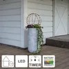 A decorative sphere light on a planter with plants, placed on a wooden deck in front of a white building. Produktbild-Vorschau