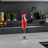 A red Bamix hand blender stands on a light gray countertop in a modern kitchen. The background features dark gray cabinets, a black backsplash, and a black faucet over a sink. Potted plants and jars of pasta are visible on a shelf behind the blender. Voir les images du produit