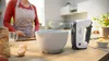 A person in an apron is preparing to use a hand mixer in a kitchen. Produktbild-Vorschau