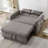 A grey sofa bed is shown in a living room setting, with a small side table and lamp to the left, and books to the right. Náhled obrázku produktu