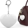 A white heart-shaped keychain is shown on the left, and a similar keychain is attached to a brown handbag on the right. Produktbild-Vorschau