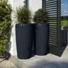 Two dark gray rain barrels are placed on a patio next to a wall with a vine, with a wooden screen in the background. Produktbild-Vorschau