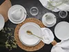 A flat lay of a white porcelain dinnerware set arranged on a dark surface, with a woven placemat and decorative dried flowers. Náhľad obrázku produktu