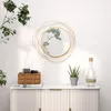 A round mirror with a gold metal frame is mounted on a white wall above a white cabinet. The cabinet holds a woven basket with items, a stack of books, a potted plant, and a clear glass vase. A branch with green leaves hangs from the upper left. Náhľad obrázku produktu