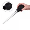 A hand is holding a black and clear turkey baster with a cleaning brush in the background. Produktbild-Vorschau