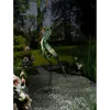 A decorative metal bird sculpture, resembling a peacock or flamingo, stands on thin legs in a garden setting with gravel and flowers. Produktbild-Vorschau