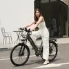 A woman is sitting on an electric bike in front of a building with outdoor seating. Produktbild-Vorschau