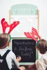 Two children wearing reindeer antlers are playing in front of a small wooden theater stage. One child is drawing on a chalkboard attached to the stage. Produktbild-Vorschau