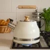 A cream-colored kettle with a wooden handle sits on a gas stove with a blue flame underneath. In the background, a basket of croissants and a French press are visible. Náhled obrázku produktu