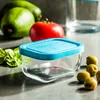A small glass container with a blue lid is placed on a gray surface, surrounded by fresh ingredients like olives, basil, tomatoes, and limes. Produktbild-Vorschau