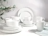 A collection of white porcelain dinnerware, including plates, bowls, and mugs, arranged on a white surface with a sprig of greenery in the background. Produktbild-Vorschau