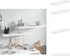 Two white floating shelves mounted on a light gray wall, decorated with various items including bottles, a box, and books. A desk with art supplies and magazines is visible below. Náhľad obrázku produktu