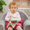 A young child sits in a gray armchair holding a colorful fabric book. Voir les images du produit