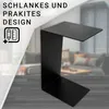 A black metal side table with a C-shaped design is placed on a wooden floor in front of a blurred living room setting with a sofa and coffee table. Produktbild-Vorschau