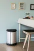 A silver metallic trash can with a black lid and foot pedal is placed on a wooden floor next to a table with a white tablecloth. There are framed pictures on the light blue wall behind the table. Anteprima dell’immagine prodotto