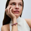 A woman wearing jewelry, including rings and a charm bracelet, with her hand resting on her chin. Anteprima dell’immagine prodotto