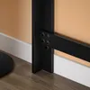A close-up view of the black metal frame of a table, showing the connection points with bolts, against a wall with white baseboard and wooden floor. Produktbild-Vorschau