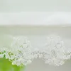 A close-up of white lace embroidery on a sheer fabric, with a blurred green background. Produktbild-Vorschau