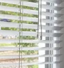 A close-up view of white horizontal blinds with green foliage visible through the slats. Produktbild-Vorschau