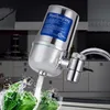 A water filter attached to a kitchen faucet, with water spraying onto fresh green vegetables in a sink. Produktbild-Vorschau